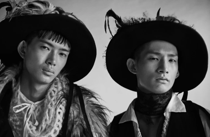 Two male models in wide-brimmed hats with feathers and textured layered garments in a black and white portrait.