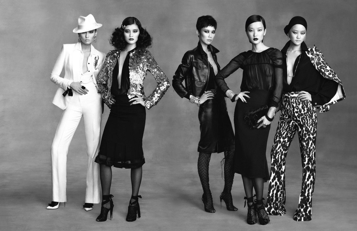Five fashion models wearing designer suits and dresses in a studio shot. Black and white image of women in elegant attire.