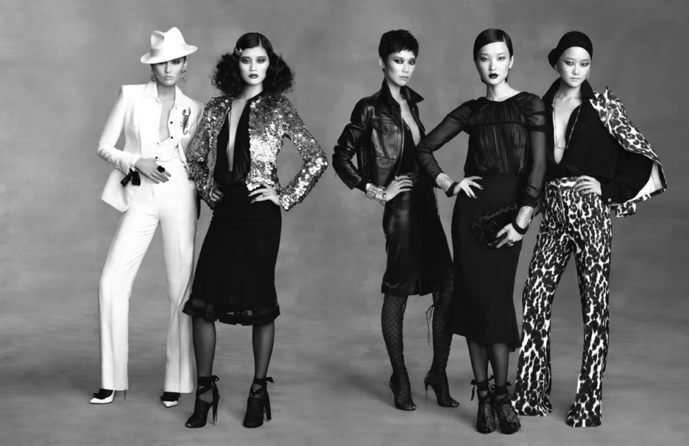 Five fashion models wearing designer suits and dresses in a studio shot. Black and white image of women in elegant attire.