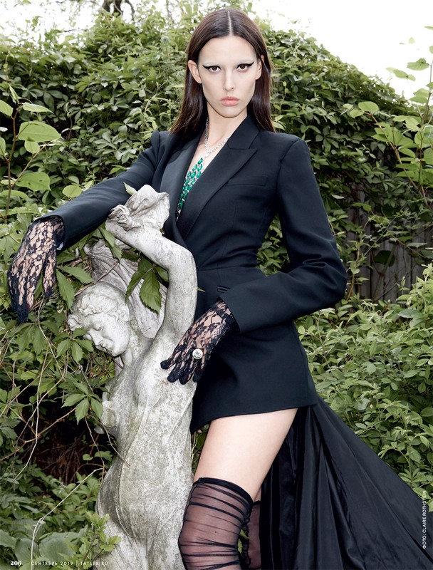 A woman in a black blazer, long skirt, and lace gloves poses next to a stone statue in a lush green garden setting with natural light.