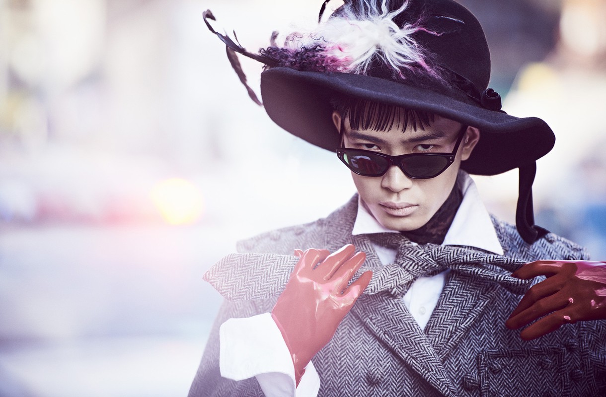 Person in a wide-brimmed hat and sunglasses poses in a herringbone jacket with red gloves, creating a high-fashion editorial image.