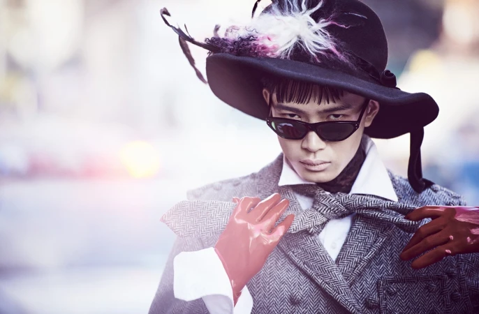 Person in a wide-brimmed hat and sunglasses poses in a herringbone jacket with red gloves, creating a high-fashion editorial image.