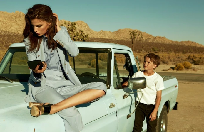 A woman sits on the hood of a vintage pickup truck in the desert looking at a compact mirror while a young boy looks on from inside.