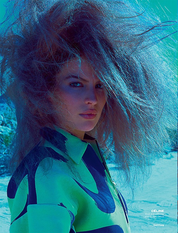 Close-up of a fashion model with voluminous blue-grey hair, wearing a green and black patterned shirt in an outdoor environment.