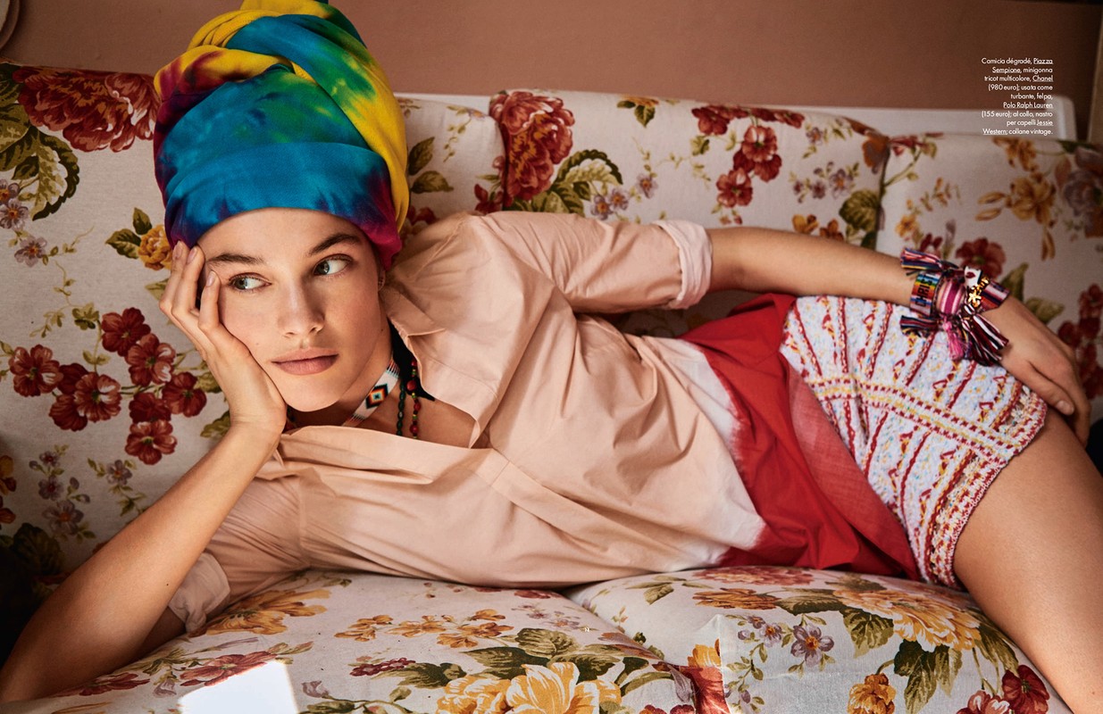 Woman lies on a floral sofa wearing a colorful headscarf, blouse, and patterned shorts looking slightly left in an indoor setting.