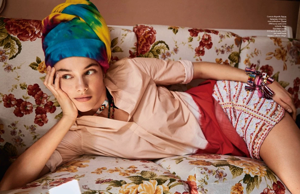 Woman lies on a floral sofa wearing a colorful headscarf, blouse, and patterned shorts looking slightly left in an indoor setting.