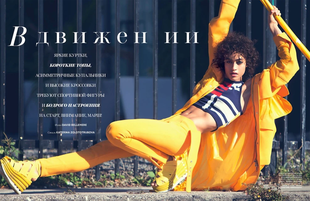 Fashion editorial shot of a woman in a yellow jacket and pants leaning against a gray barrier outdoors with large text overlay.