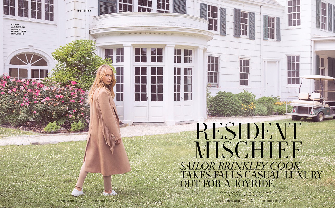 Woman poses in a tan coat and pants on a lawn in front of a white house with green shutters and a golf cart on a sunny day.