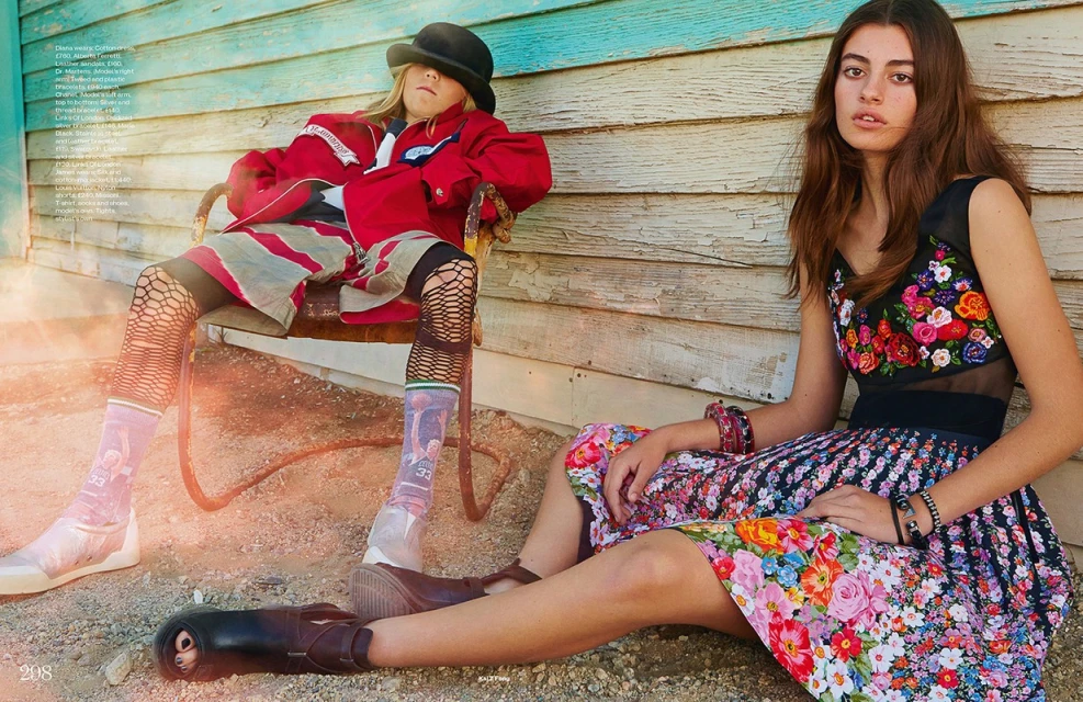 Two young adults sit outdoors against a weathered blue wall. One wears a red jacket and a bowler hat, the other a floral dress.