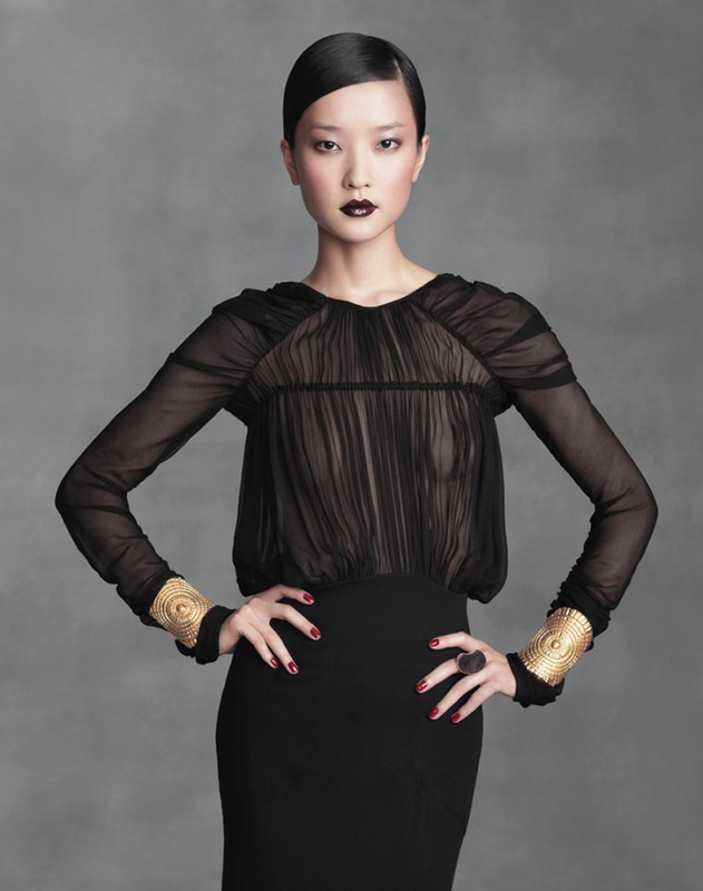Elegant Asian woman in a black dress with long sleeves and gold cuff bracelets, posing against a neutral gray backdrop in a studio shot.