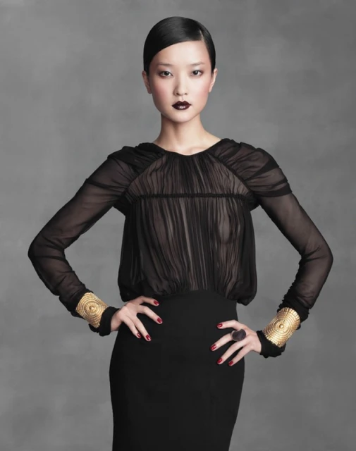 Elegant Asian woman in a black dress with long sleeves and gold cuff bracelets, posing against a neutral gray backdrop in a studio shot.