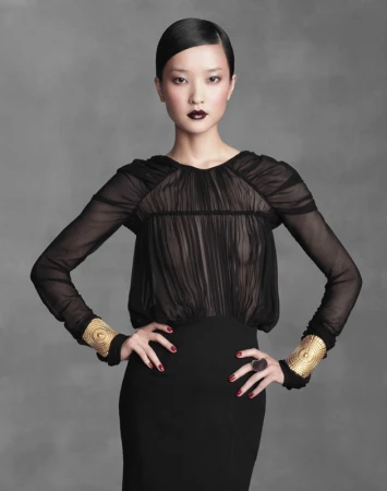 Elegant Asian woman in a black dress with long sleeves and gold cuff bracelets, posing against a neutral gray backdrop in a studio shot.
