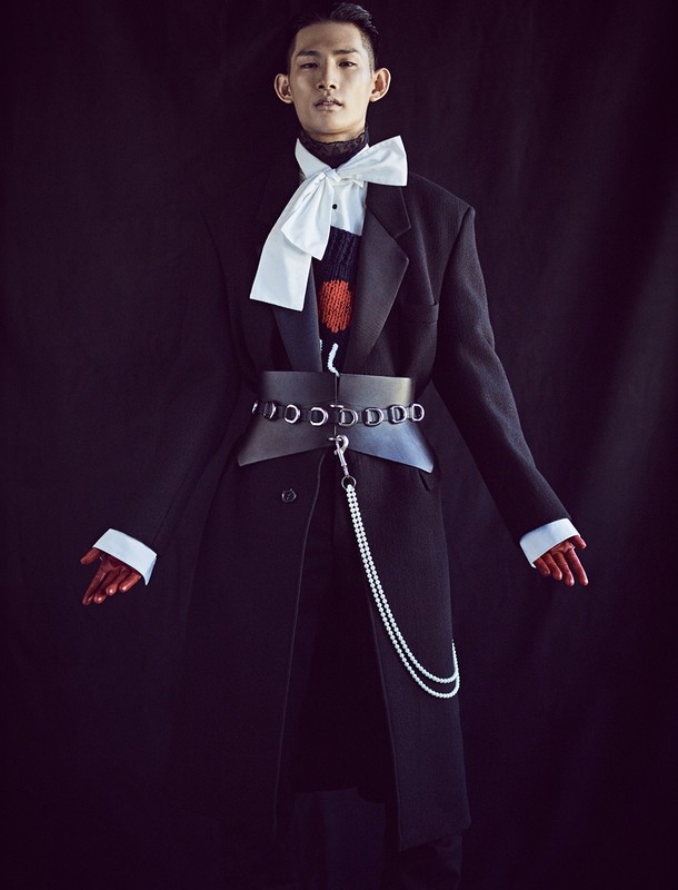 Stylish Asian male in an oversized black coat, accessorized with a large bow tie, wide belt, and pearl chain, set against a dark backdrop.