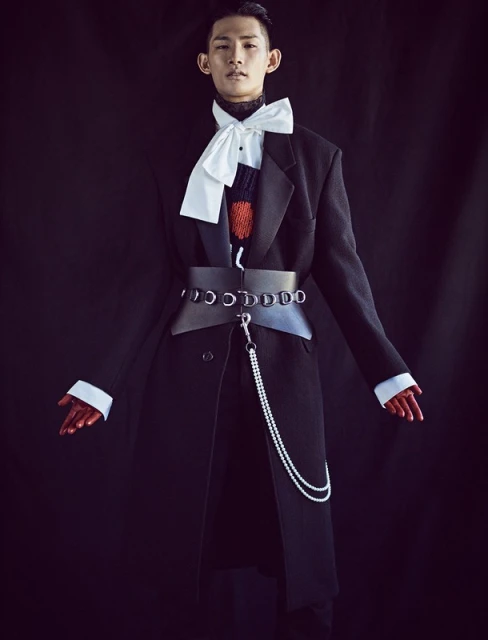 Stylish Asian male in an oversized black coat, accessorized with a large bow tie, wide belt, and pearl chain, set against a dark backdrop.