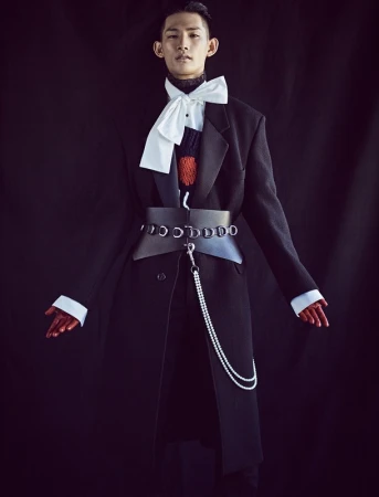 Stylish Asian male in an oversized black coat, accessorized with a large bow tie, wide belt, and pearl chain, set against a dark backdrop.