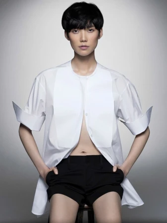 Model with short black hair wearing an unbuttoned white shirt and black shorts against grey backdrop