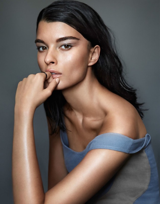 Close-up portrait of a woman with dark hair and light eyes, wearing a blue top, posing in front of a gray background.