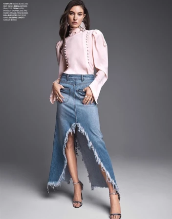 Model in pink high-neck blouse and high-low denim skirt posing against grey background with makeup by Chris Colbeck