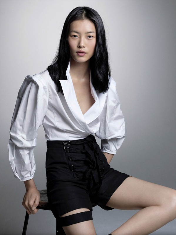 An Asian woman sits on a stool in a studio. She wears a white blouse with black details and high-waisted black shorts with corset lacing.