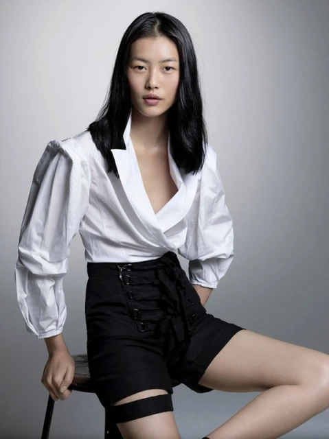 An Asian woman sits on a stool in a studio. She wears a white blouse with black details and high-waisted black shorts with corset lacing.