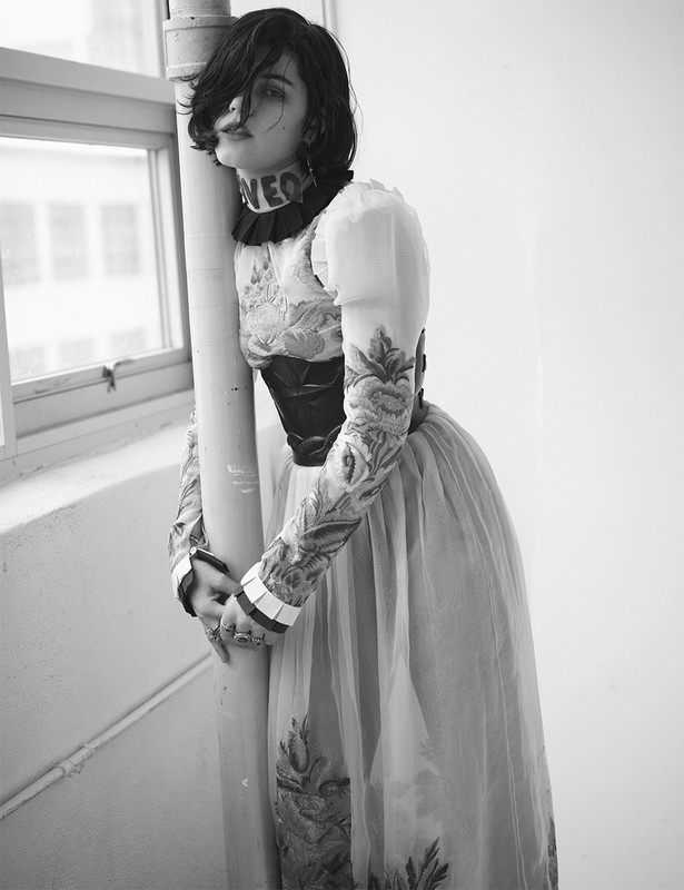 Black and white portrait of a woman with dark hair in a floral dress leaning on a pipe indoors, looking up towards a window.