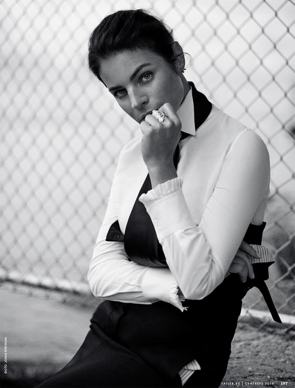Black and white portrait of a woman in a white collared shirt and dark tie leaning against a chain-link fence.