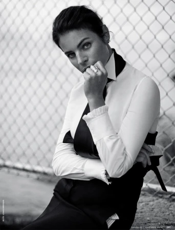 Black and white portrait of a woman in a white collared shirt and dark tie leaning against a chain-link fence.