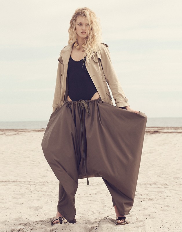 Adult female model in a black bodysuit, trench coat, and brown harem pants poses on a beach with a neutral sky and sand background.