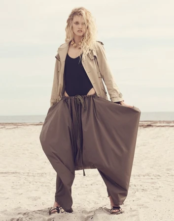 Adult female model in a black bodysuit, trench coat, and brown harem pants poses on a beach with a neutral sky and sand background.