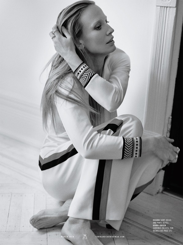 Black-and-white portrait of a blonde woman in a white tracksuit with dark trim, sitting on a floor against a neutral background.