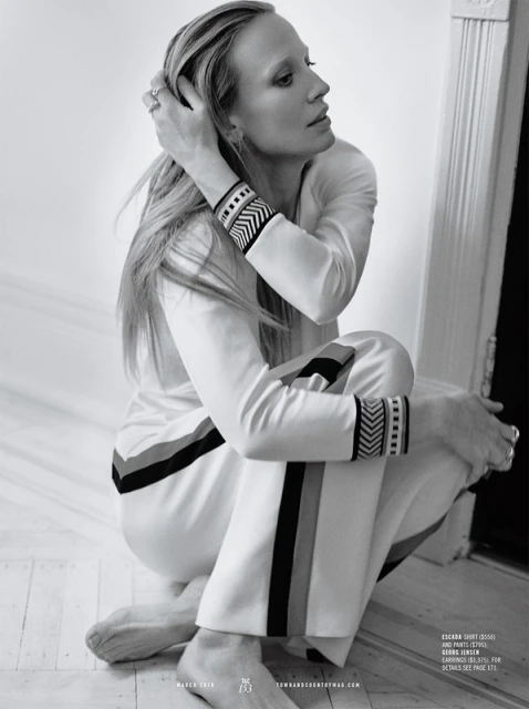 Black-and-white portrait of a blonde woman in a white tracksuit with dark trim, sitting on a floor against a neutral background.