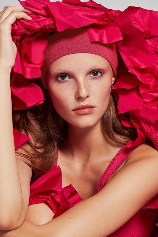 Young woman in a fashion portrait, wearing a matching rose colored head covering and dress, surrounded by large rose-colored ruffles.