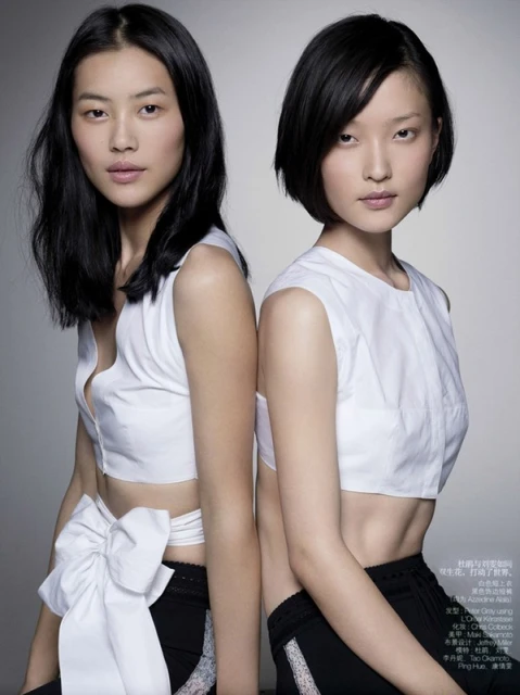 Two Asian women pose back-to-back in white crop tops and black bottoms against a gray background in a beauty and fashion editorial shot.