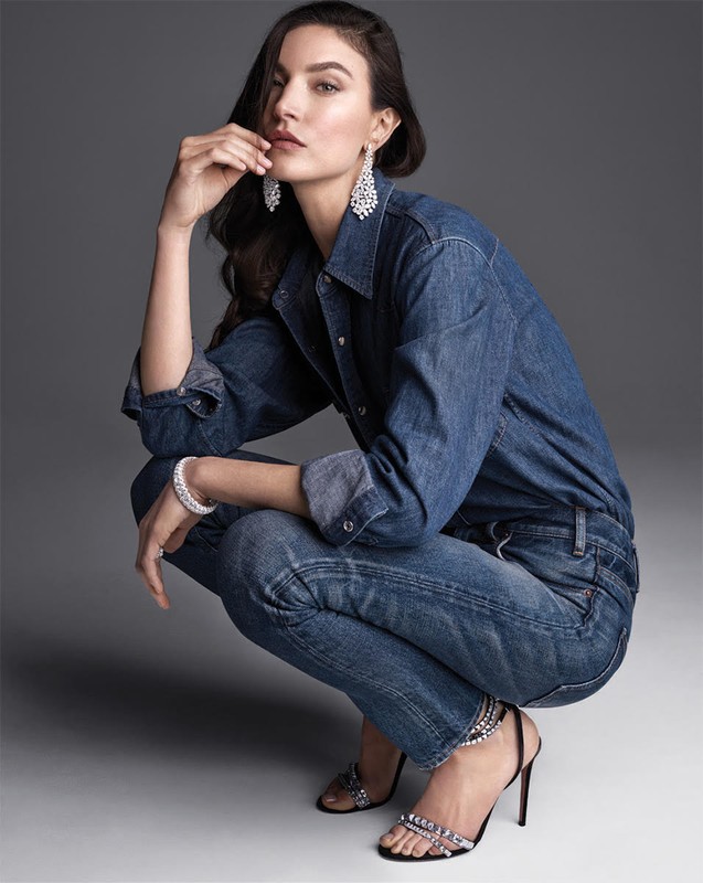 Portrait of an adult female model in a denim outfit, crouching in a photo studio, wearing chandelier earrings and jeweled high heels.