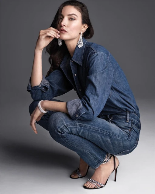 Portrait of an adult female model in a denim outfit, crouching in a photo studio, wearing chandelier earrings and jeweled high heels.