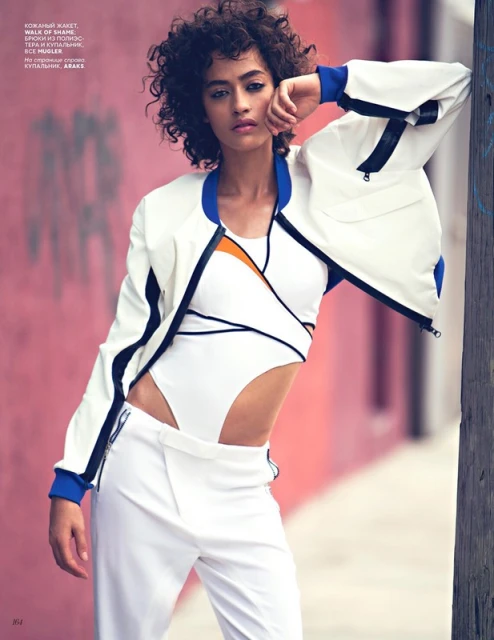 A woman poses in a white jacket and pants against a pink wall, looking at the camera, displaying a modern, fashionable style outdoors.