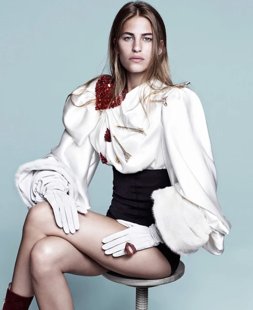 Fashion shot of a fair-skinned woman with long brown hair, wearing a white, decorative jacket and black shorts, seated on a stool.