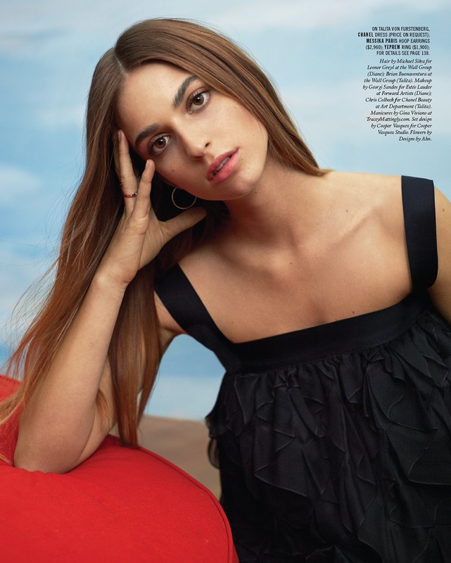 Portrait of a woman with long brown hair wearing a black dress against a sky background. She is posing with hand near her face.