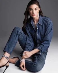 Model wearing a blue denim shirt and matching jeans seated against a grey gradient background with diamond jewellery