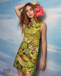 Woman in gold and green patterned sleeveless dress holding a large pink flower against a blue painted wall