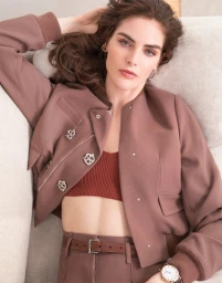 Woman wearing a brown jacket with silver buttons and a reddish-brown crop top, reclining on a beige sofa.