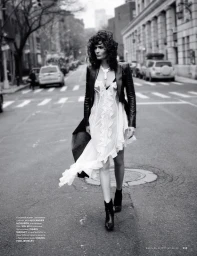 Black and white editorial portrait of a woman walking down a city street wearing a white ruffled dress and dark jacket.