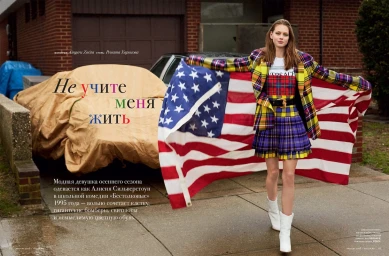 Model holding a large American flag wearing a yellow plaid blazer and matching plaid skirt standing on a driveway in Los Angeles.