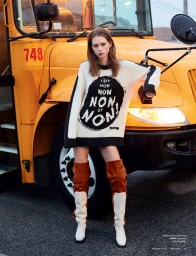 Model wearing printed sweater and tall boots leaning against a large yellow school bus in Los Angeles
