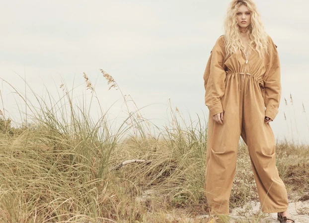 Blonde female model wearing a camel jumpsuit standing amongst tall coastal dune grasses