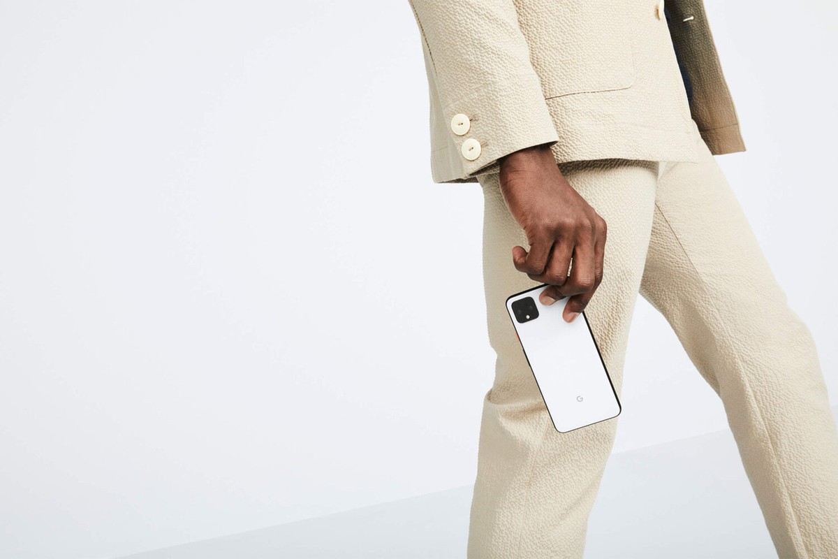 Person in a beige suit holding a white smartphone against a white background in a studio setting, capturing a minimalistic tech fashion aesthetic.