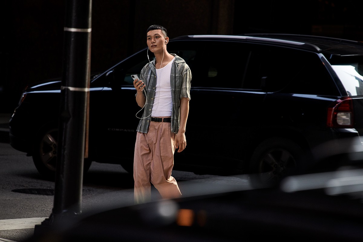 Young man with headphones uses a smartphone on a city street, wearing casual attire, with a black car parked in the background.