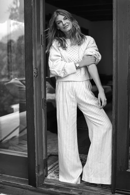 Blonde woman in a cable knit sweater and striped palazzo pants stands in a doorway with light coming through, monochrome.