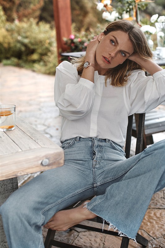 A fair-skinned woman with freckles poses outdoors in a white blouse and jeans, seated at a wooden table with a drink, in soft, natural light.