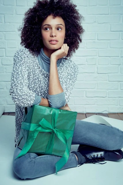 Young woman with afro sits cross-legged indoors against a brick wall, holding a green, ribbon-wrapped gift box in her lap.